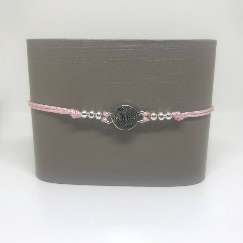 May include: A pink string bracelet with silver beads and a silver compass charm. The compass charm has the letters N, S, E, and W engraved on it.