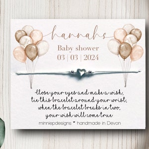 May include: A white card with a watercolour illustration of balloons in shades of brown and pink. The card says "hannahs Baby shower 03|03|2024" and includes a white string bracelet with a silver heart charm. The text below the bracelet reads "Close your eyes and make a wish, tie this bracelet around your wrist, when the bracelet breaks in two, your wish will come true. minniepdesigns handmade in Devon."