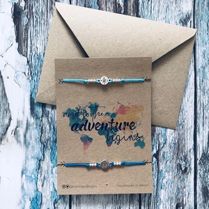 May include: Two blue friendship bracelets with silver compass charms on a brown card with the text "and so the adventure begins." The card also has a watercolour world map design and the text "@tinniedpdesigns handmade in devon".