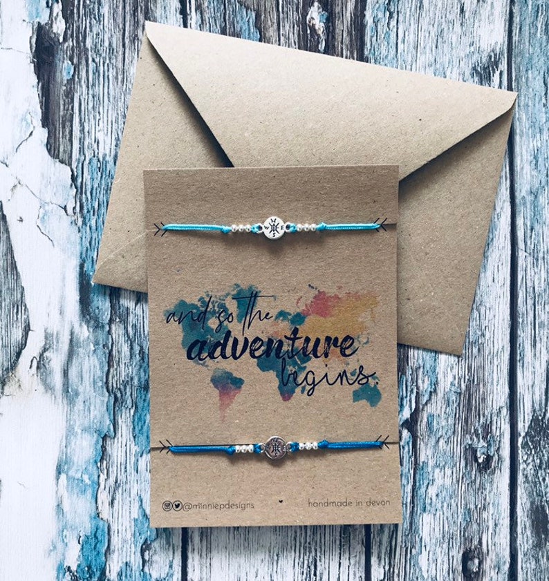 May include: Two blue friendship bracelets with silver compass charms on a brown card with the text "and so the adventure begins." The card also has a watercolour world map design and the text "@tinniedpdesigns handmade in devon".