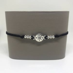 May include: A black cord bracelet with silver beads and a compass charm. The compass charm has the directions N, S, E, and W engraved on it.