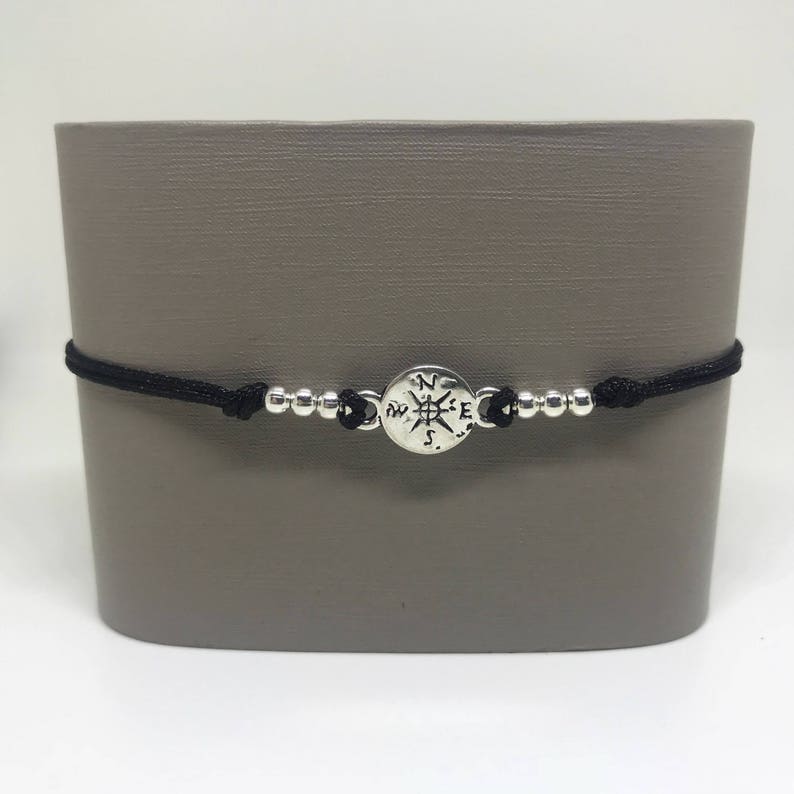 May include: A black cord bracelet with silver beads and a compass charm. The compass charm has the directions N, S, E, and W engraved on it.