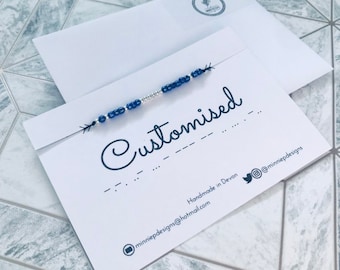 May include: A white card with the word "Customised" written in black cursive. The card also includes the text "Handmade in Devon" and an email address and Twitter handle for the business.