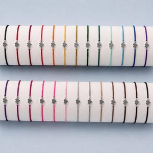 May include: A collection of twenty-six thin cord bracelets displayed on two white cylindrical holders. Each bracelet features a small silver-toned heart charm flanked by two tiny silver beads, available in a variety of solid colours including black, red, orange, yellow, green, blue, purple, pink, grey, beige, and brown.