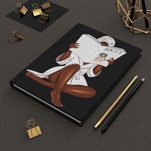 Op de afbeelding: Een zwart notitieboek met een witte illustratie van een persoon in een witte badjas die een tijdschrift leest met het woord "Vogue" op de omslag.