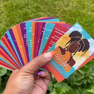 May include: A deck of affirmation cards with the title "Victory Affirmation Cards" and a colorful illustration of a woman with curly hair. The cards are fanned out in a hand.