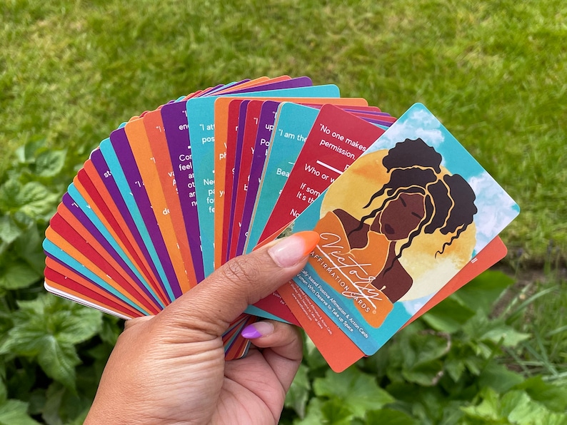 May include: A deck of affirmation cards with the title "Victory Affirmation Cards" and a colorful illustration of a woman with curly hair. The cards are fanned out in a hand.