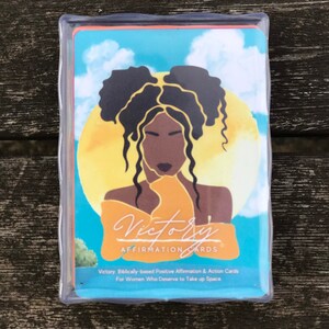 May include: A deck of affirmation cards with a blue background and a yellow sun. The cards feature a black woman with her hair in two buns. The text on the cards reads "Victory Affirmation Cards" and "Victory: Biblically-based Positive Affirmation & Action Cards For Women Who Deserve to Take up Space."