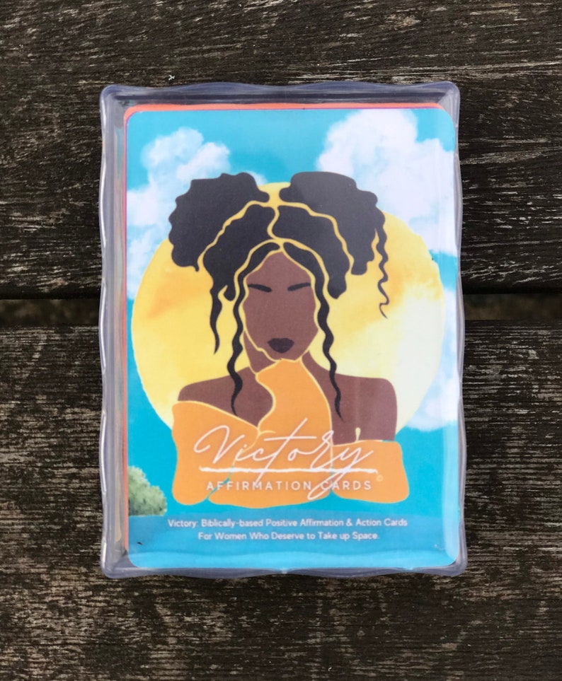May include: A deck of affirmation cards with a blue background and a yellow sun. The cards feature a black woman with her hair in two buns. The text on the cards reads "Victory Affirmation Cards" and "Victory: Biblically-based Positive Affirmation & Action Cards For Women Who Deserve to Take up Space."