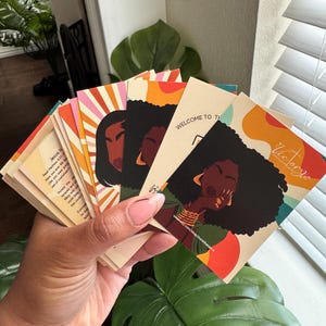 May include: A hand holds a stack of colorful cards with illustrations of Black women and abstract designs. The cards feature text, including "WELCOME TO THE" and "Victory She". The color palette includes orange, red, and brown.
