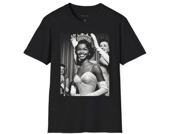 May include: Black and white photograph of a woman wearing a crown and a white dress. She is smiling and holding a microphone. The photo is printed on a black t-shirt.