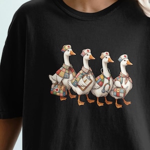 Puede incluir: Camiseta negra con una ilustración caprichosa de cuatro gansos con sombreros y chalecos. Los gansos están representados en fila, cada uno con patrones únicos y accesorios médicos. El diseño está centrado en la camiseta.