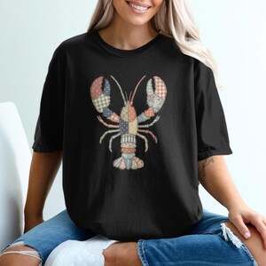 Puede incluir: Camiseta negra con un diseño de langosta de patchwork. La langosta está hecha de varios patrones y colores de tela, incluyendo rojo, azul y verde. La camiseta es de estilo casual y relajado.
