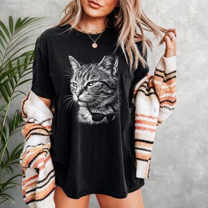 May include: Black t-shirt with a detailed grayscale illustration of a cat wearing a bow tie. The cat's face is the focal point, with its eyes and whiskers clearly visible. The shirt is worn with a striped sweater.