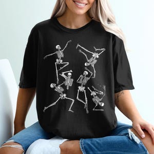 Może przedstawiać: Czarny t-shirt z białym nadrukiem tańczących szkieletów w różnych pozach. Szkielety przedstawione są w żywej i dynamicznej aranżacji, z wyciągniętymi ramionami i nogami. Koszulka jest w stylu casual i wygodnym.