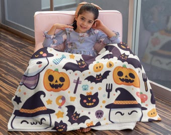 Op de afbeelding: Een Halloween-deken met een herhalend patroon van pompoenen, vleermuizen, heksenhoeden, geesten en sterren in oranje, zwart, paars en wit. De deken is gedrapeerd over een persoon die in een roze stoel zit.