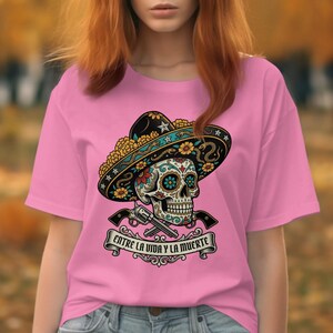 May include: Pink t-shirt featuring a colorful sugar skull design wearing a sombrero, with crossed pistols and the text "Entre la Vida y la Muerte". The design is in black, yellow, blue, and green.