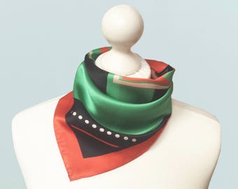 May include: A square silk scarf with a geometric design in green, black, and red. The scarf is displayed on a white mannequin head. The design includes white dots and lines.