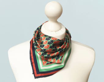 May include: A square scarf with a repeating fan-like pattern in red, navy, and green on a white background. The scarf is displayed on a white mannequin. The scarf has a navy, red, and green border.