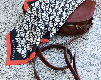 May include: A brown leather crossbody bag with a gold zipper and strap, paired with a patterned silk scarf. The scarf features a black and white floral design with a red border. The scarf has the text "CINDA CLARK".