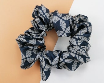 May include: A navy blue and gray patterned scrunchie. The fabric features a repeating geometric design. The scrunchie is gathered and elasticized, designed to hold hair. The background is split between a peach and white color.