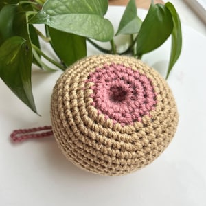 May include: A round, crocheted object featuring a tan-colored base with a circular, textured pink center. The item is crafted from yarn with visible stitches and includes a small, thin pink loop extending from the side of the main spherical form.
