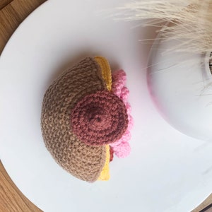 May include: A crocheted breast model featuring a light brown skin tone, a dark brown circular areola, and a raised nipple. The side profile reveals a yellow layer and a cluster of small, textured pink spheres representing mammary tissue.