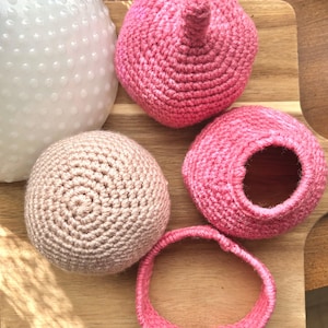 May include: A set of crocheted items including a solid pink sphere with a small stem, a solid beige sphere, a hollow pink bowl-like shape, and a separate pink circular ring. The items are crafted with a visible yarn texture and consistent stitch pattern.
