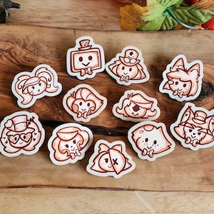 May include: A collection of eleven die-cut stickers featuring cartoon characters with various hairstyles and accessories. The stickers are outlined in brown and have a white background. The stickers are arranged on a wooden surface.