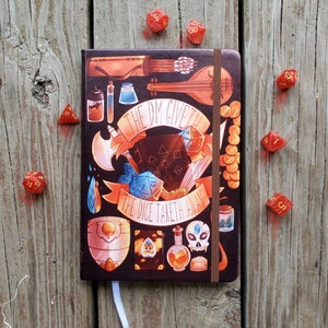 May include: A brown leather-bound journal with a colorful illustration of dice, a sword, and a shield. The text "The DM Giveth The Dice Taketh Away" is printed on the cover.