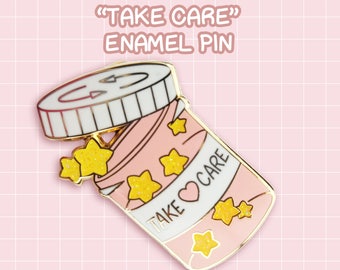 Op de afbeelding: Roze emaille pin in de vorm van een pillenfles met een wit deksel en gouden accenten. De fles is open en gevuld met gouden glittersterren. Het etiket op de fles zegt "Take Care" met een hartje. De pin is op een roze en witte geruite achtergrond.