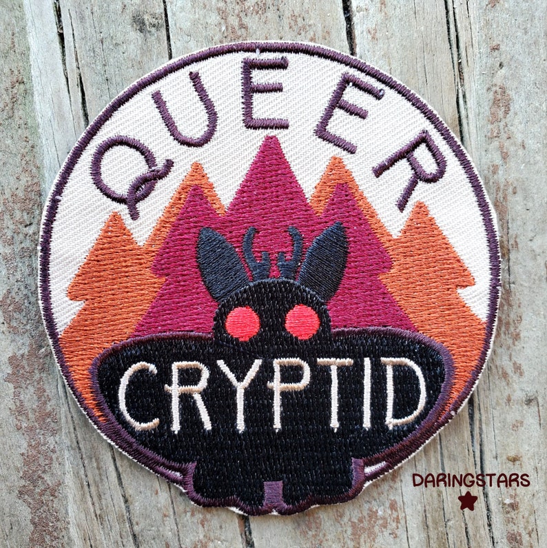 May include: A round embroidered patch with the text "Queer Cryptid" in white lettering on a black background. The patch features a black moth with red eyes in front of a stylised mountain range with orange and purple trees.