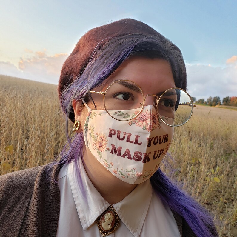 May include: A person wearing a brown beret and round glasses with a white face mask with floral print and the text "PULL YOUR MASK UP" in red.