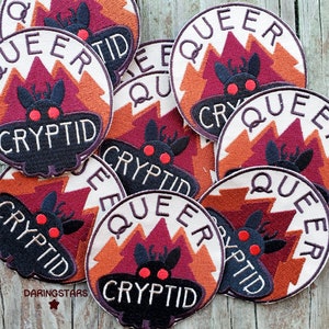 May include: Eight embroidered patches with a white background and red, orange, and brown accents. Each patch features a black moth with red eyes and the text "Queer Cryptid".