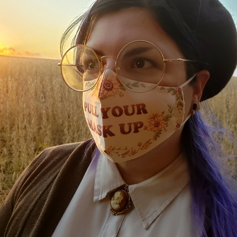 May include: A person wearing a face mask with the text "Pull Your Mask Up" printed on it. The mask is white with floral designs and the text is in brown and red. The person is wearing a brown jacket and a white shirt with a cameo brooch.