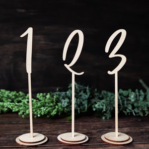 Peut inclure: Trois numéros de table en bois avec les chiffres 1, 2 et 3 écrits en cursive. Les numéros sont fixés à des bâtonnets en bois avec des bases rondes en bois.