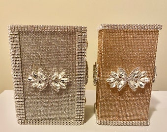 May include: Two decorative boxes covered in rhinestones. One box is silver and the other is gold. Both boxes have a rhinestone bow on the front.