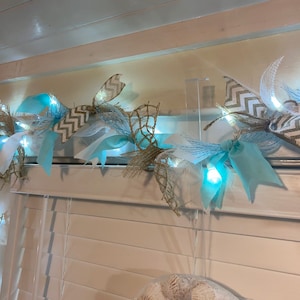 May include: A decorative garland featuring a combination of light blue, white, and tan ribbons, accented with small white lights. The garland includes chevron patterned ribbons and a net-like design, creating a coastal or nautical theme.