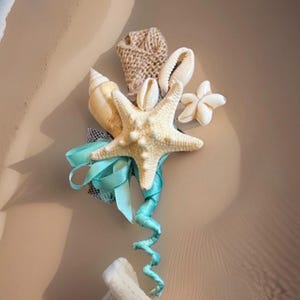 May include: A decorative boutonniere featuring a central cream-colored starfish, a spiral seashell, a small white starfish, and a cowrie shell. The arrangement is accented with a textured tan burlap rose, a turquoise satin ribbon, and netting.