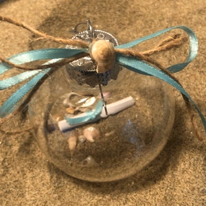 May include: A clear glass ornament with a blue ribbon and twine bow. The ornament contains seashells and sand.