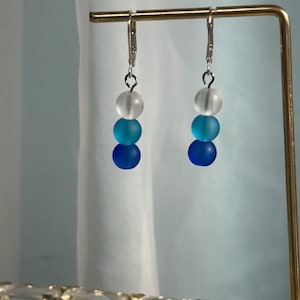 May include: A pair of drop earrings featuring three stacked frosted beads in a gradient color scheme. The beads transition from a translucent white at the top to a bright turquoise in the middle and a deep royal blue at the bottom, suspended from silver-toned metal leverback hooks.