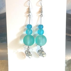 May include: A pair of dangle earrings featuring a beach theme. Each earring has a silver-tone fish hook, a series of blue and turquoise beads, and a silver-tone charm shaped like a sand dollar. The earrings are displayed on a white card.