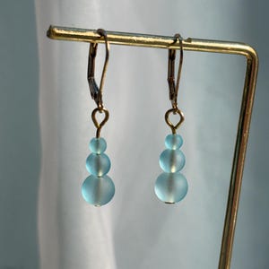 May include: A pair of drop earrings featuring three graduated frosted light blue glass beads suspended from gold-toned metal leverback hooks. The beads are arranged in a vertical line, transitioning from the smallest at the top to the largest at the base.