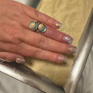 May include: A silver ring with two round, seashell-adorned settings. The hand also displays manicured nails with glitter and seashell designs. The ring and nails are set against a backdrop of sand and a starfish.