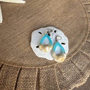 Op de afbeelding: Druppelvormige oorbellen met een lichtblauw hars- en zandontwerp, geplaatst op een witte zeester. De oorbellen hebben zilverkleurige oorhaken. De achtergrond is een jute placemat met een zeester.