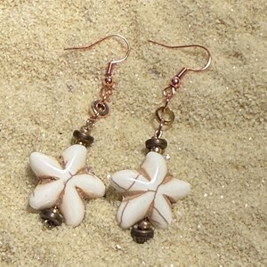 May include: A pair of dangle earrings featuring white, starfish-shaped pendants with brown accents. The earrings have copper-colored hooks and are accented with small beads. The earrings are displayed on a sandy surface.
