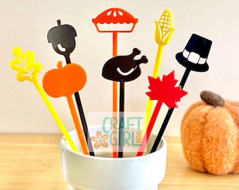 May include: A set of six colorful Thanksgiving themed acrylic drink stirrers in a white ceramic cup. The stirrers feature a variety of designs, including a turkey, a pumpkin, a cornucopia, a leaf, a pie, and a pilgrim hat.