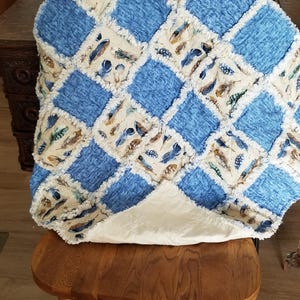 K&ouml;nnte beinhalten: Eine blaue und cremefarbene Patchwork-Decke mit Federndruck. Die Decke ist gefaltet und auf einem Holzstuhl gelegt.