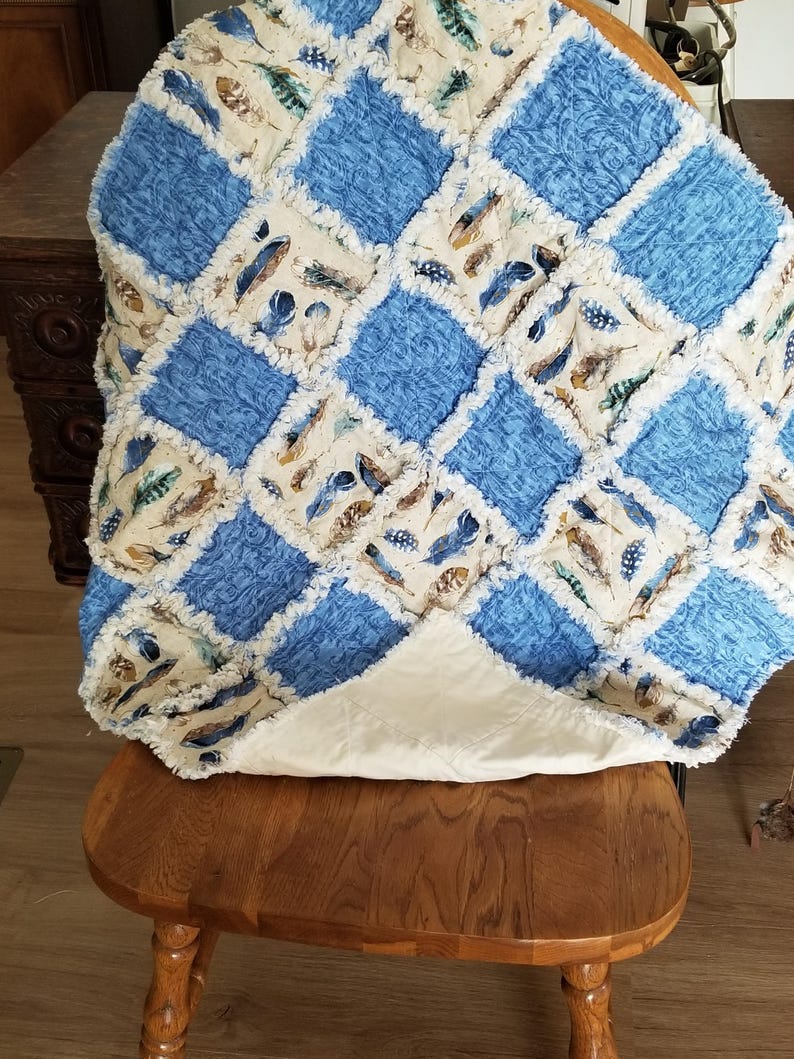 K&ouml;nnte beinhalten: Eine blaue und cremefarbene Patchwork-Decke mit Federndruck. Die Decke ist gefaltet und auf einem Holzstuhl gelegt.