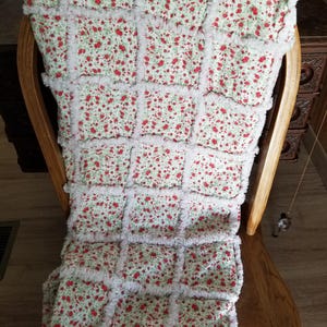 Pu&ograve; includere: Una copertina per neonati di colore bianco e rosso con stampa floreale e un bordo bianco, soffice e sfilacciato. La copertina &egrave; piegata e appoggiata su una sedia di legno.